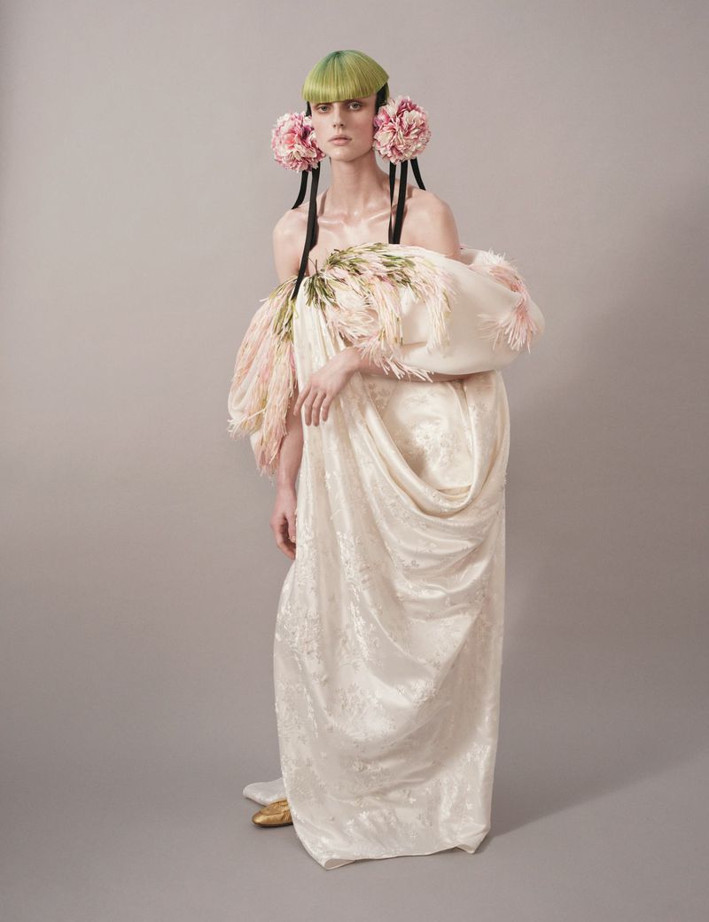 Model wears large floral decorations and elaborate draped fabric gown in a studio fashion portrait with artistic hairstyling and makeup.
