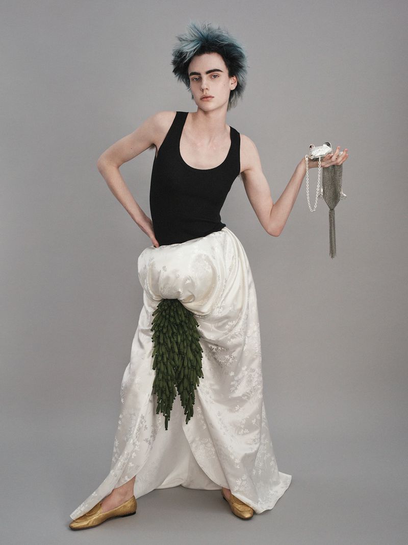 A person with spiky blue hair poses in a black tank top and a large white skirt, holding a beaded bag against a grey background.