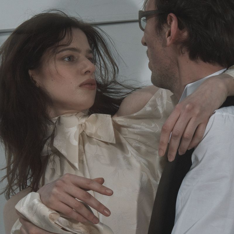 A stylish couple in close embrace, the woman in a patterned cream blouse with a bow, and the man in a shirt and tie, captured indoors.