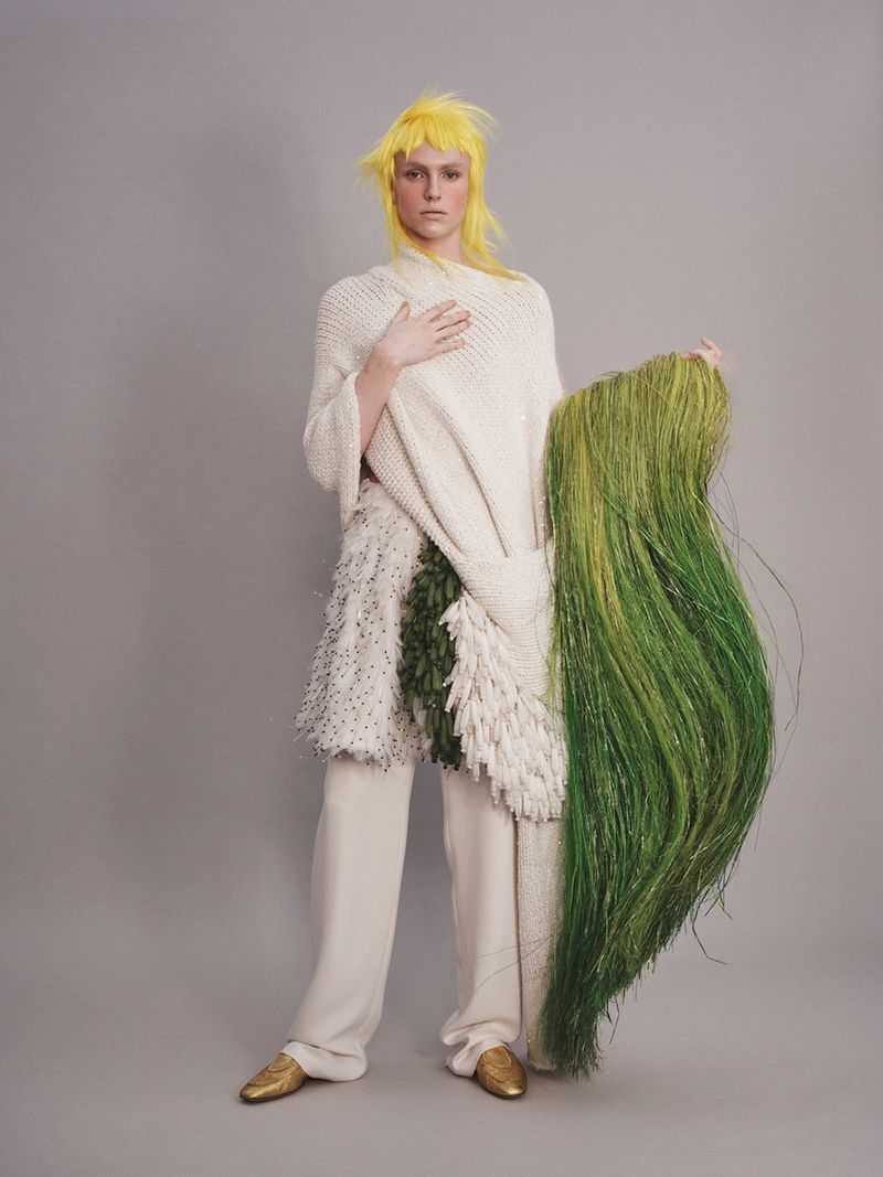Full-length portrait of a person with yellow hair wearing an off-white poncho and beige pants, holding green grasses in a photo studio setting.