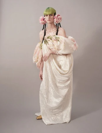 Model with green hair and floral ear decorations wearing a voluminous off-shoulder cream gown against a grey background