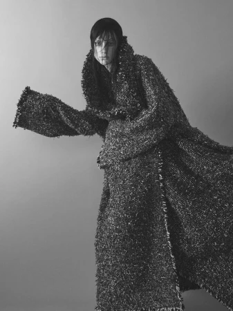 Model wearing a heavily textured dark tweed coat with wide dramatic collar against a plain grey background