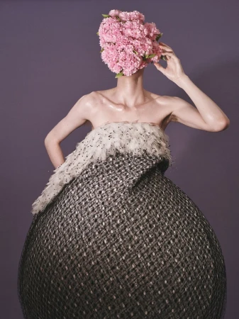 Model wearing a voluminous textured gown with head obscured by a dense cluster of pink flowers against a solid background