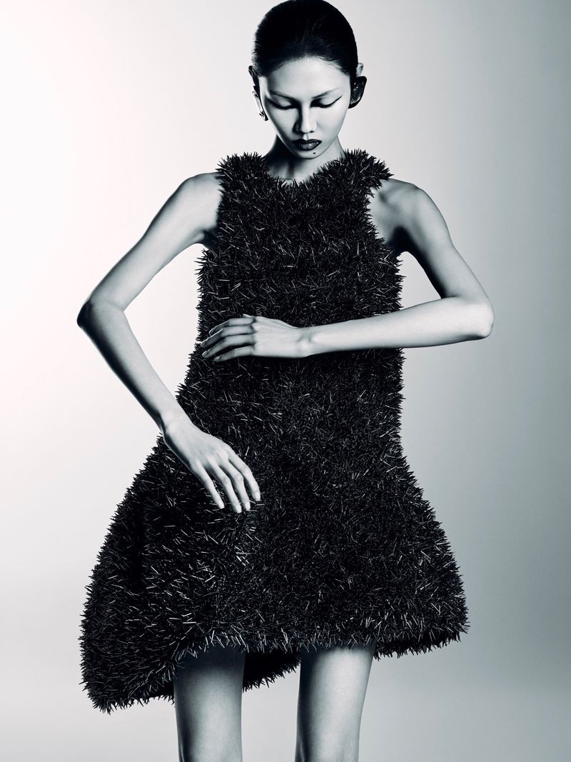 Monochromatic portrait of an Asian woman in a textured black dress, posing with hands near waist in a white studio setting.