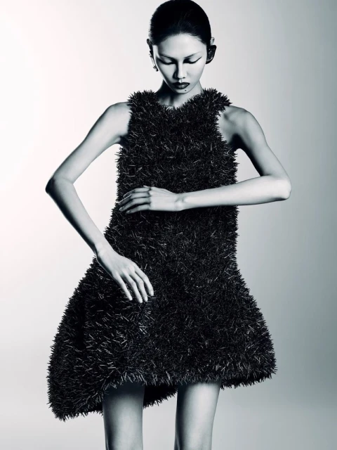 Monochromatic portrait of an Asian woman in a textured black dress, posing with hands near waist in a white studio setting.