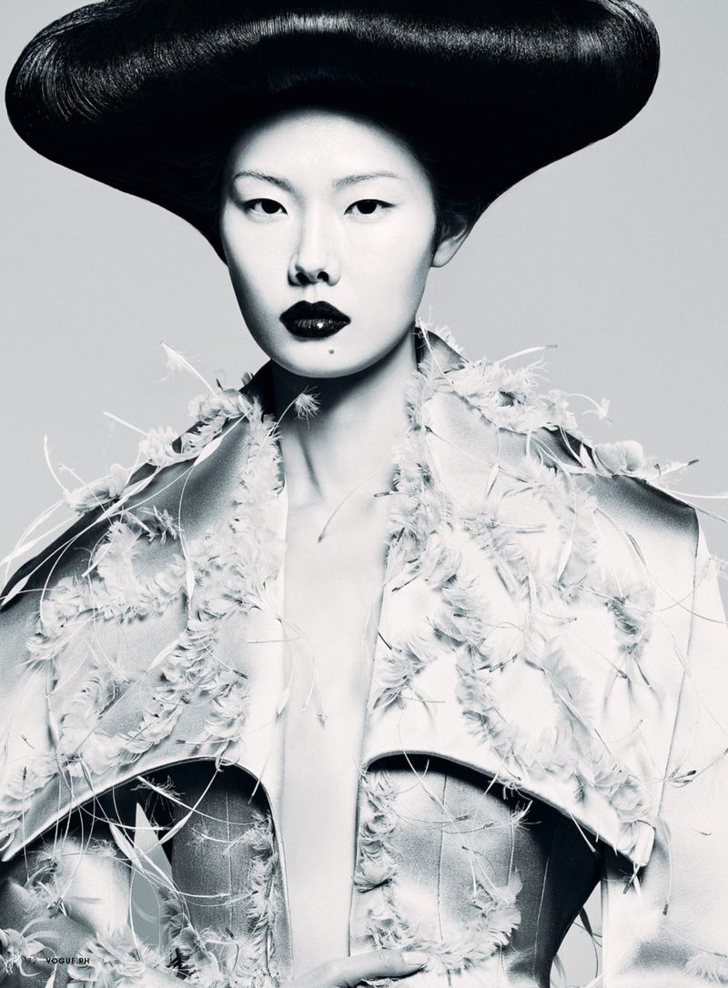 Asian woman with dramatic black hair and makeup poses in a feathered jacket against a neutral background, fashion portrait, studio lighting.