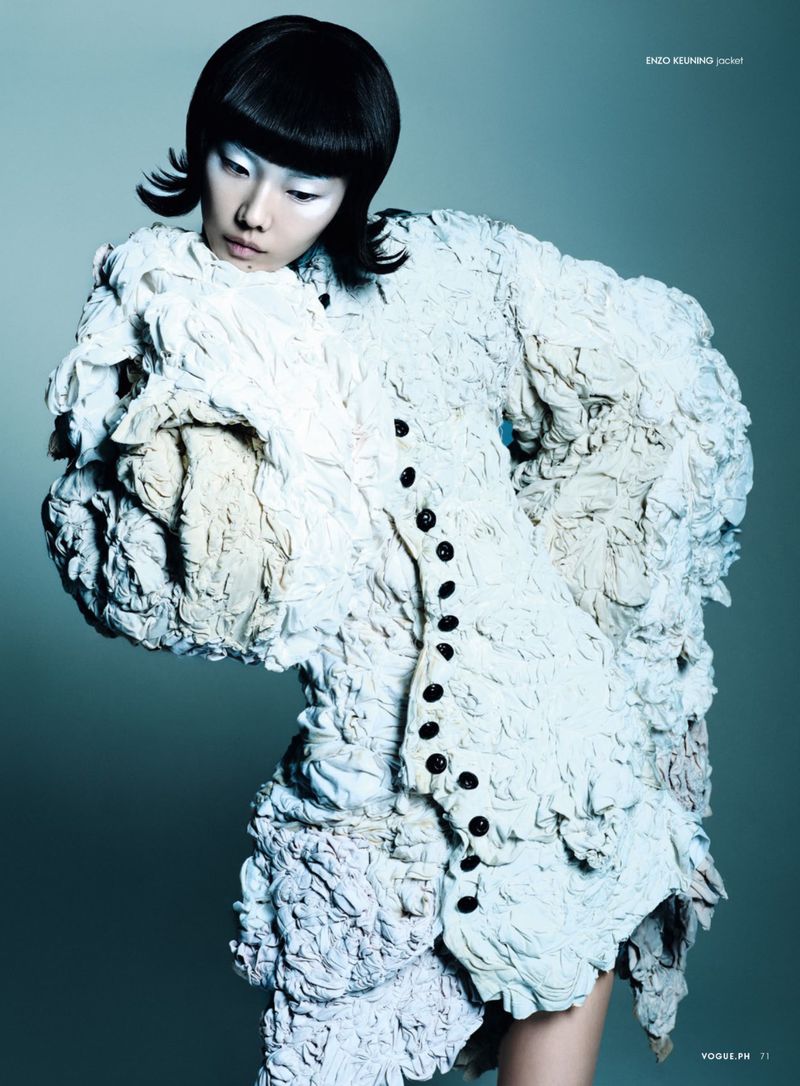 A person with bright-white skin and black hair wears a white, heavily textured jacket with oversized embellishments and dark buttons.