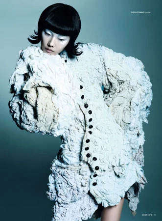 A person with bright-white skin and black hair wears a white, heavily textured jacket with oversized embellishments and dark buttons.