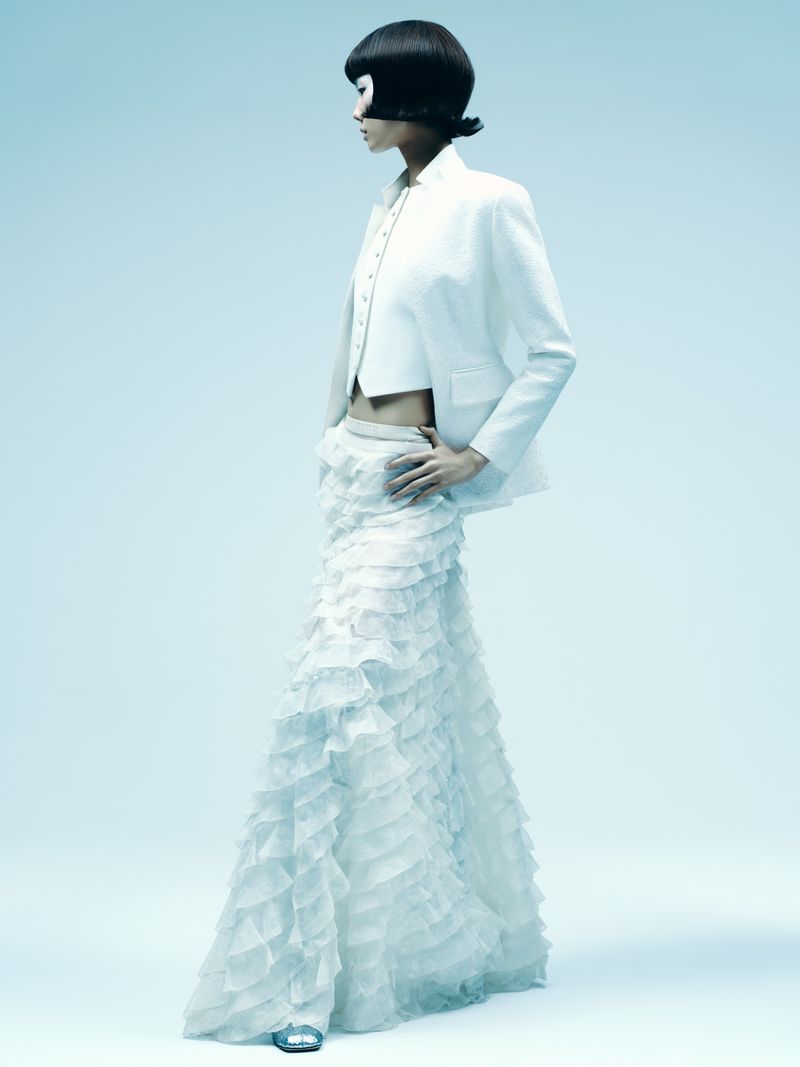 Fashion shot of an adult female with a black bob haircut wearing a white jacket, crop top, and ruffled maxi skirt in a light blue photo studio.