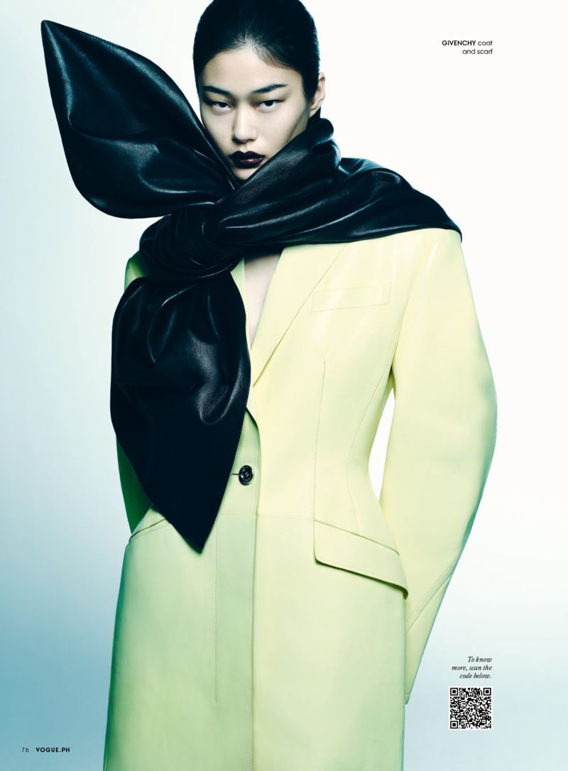 A fashionable Asian female model poses in a studio wearing a light yellow coat and a large, black, shiny bow scarf. The studio background is gradient light blue.