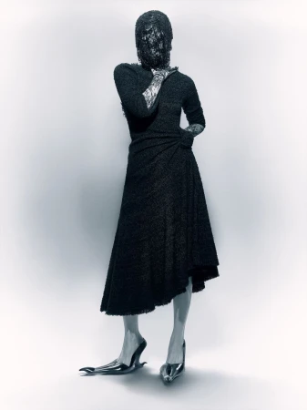 Abstract fashion image showcasing a person in a textured black dress with an obscured face and avant-garde footwear against a light background.