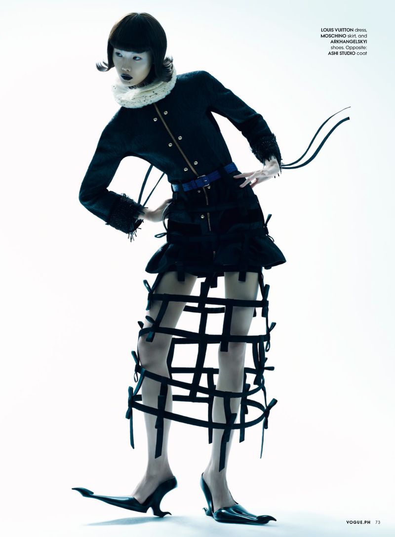 Fashion model poses in a studio wearing a black jacket, skirt with straps, statement collar, and avant-garde black heeled shoes.