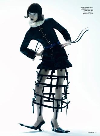 Fashion model poses in a studio wearing a black jacket, skirt with straps, statement collar, and avant-garde black heeled shoes.