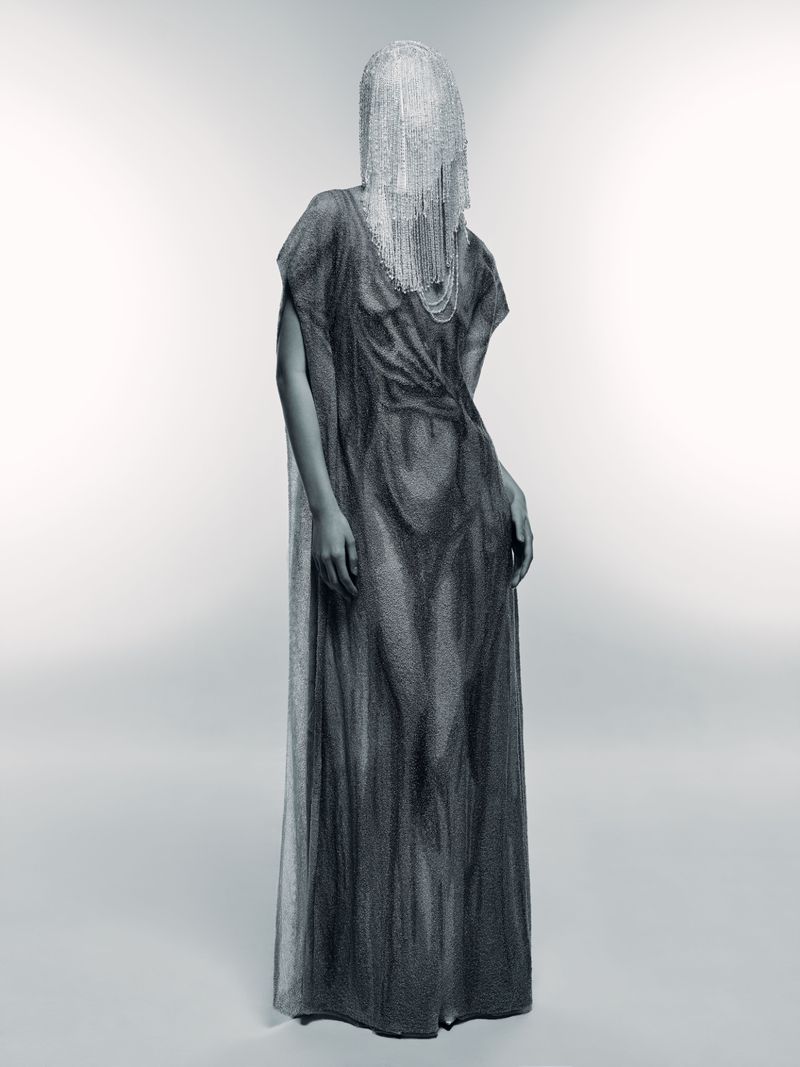 Full-length studio shot of an adult woman wearing a draped gown and face covering, posing against a gradient white background with soft lighting.