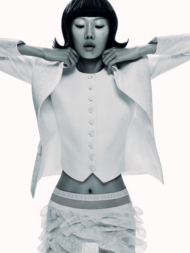 Fashion shot of an Asian woman in a white jacket and skirt. Studio lighting and neutral background. Short black bob haircut and hands near face.