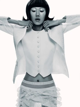 Fashion shot of an Asian woman in a white jacket and skirt. Studio lighting and neutral background. Short black bob haircut and hands near face.