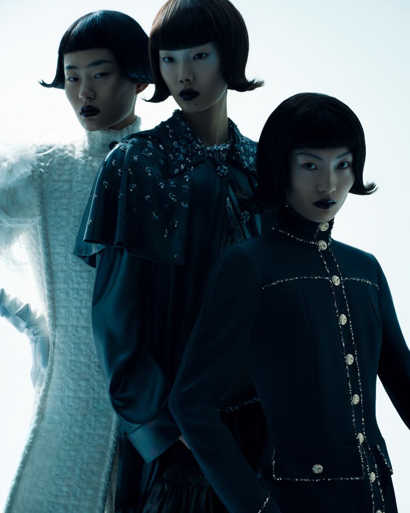 Three Asian women in avant-garde fashion pose in a studio with cool lighting. Dark lipstick and unique hairstyles add to the striking aesthetic.