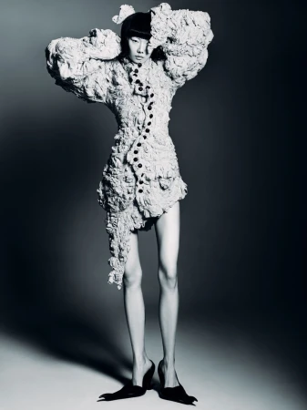 A person poses in a sculptural, textured dress and pointed heels against a neutral gray background in a studio fashion shot.