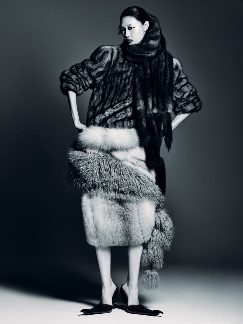 Monochrome fashion shot of a slender Asian woman in an extravagant fur outfit with a matching scarf, standing in a studio setting.