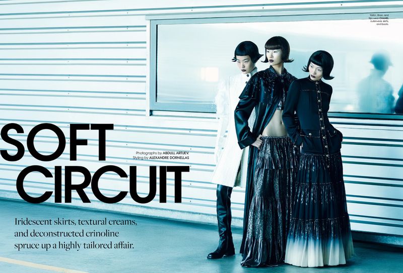 Three Asian women in modern fashion pose against a minimalist wall with a long window, showcasing avant-garde style in an indoor setting.