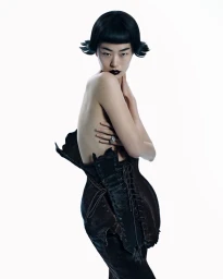 Model with dark bob haircut and dark lipstick wearing a structured laced corset against a white backdrop.