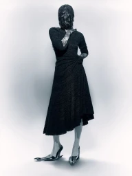 Model wearing a black dress and metallic heeled shoes with face covered by a textured mask against a grey gradient background