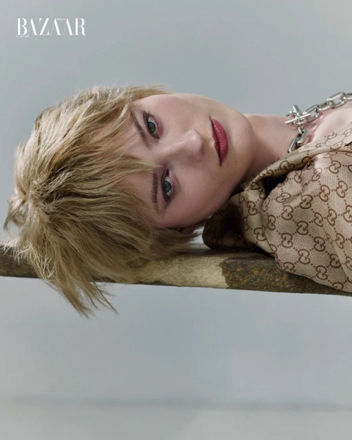 Blonde-haired model lays at an angle looking into the camera wearing an oversized chain necklace and Gucci-patterned jacket in a studio.