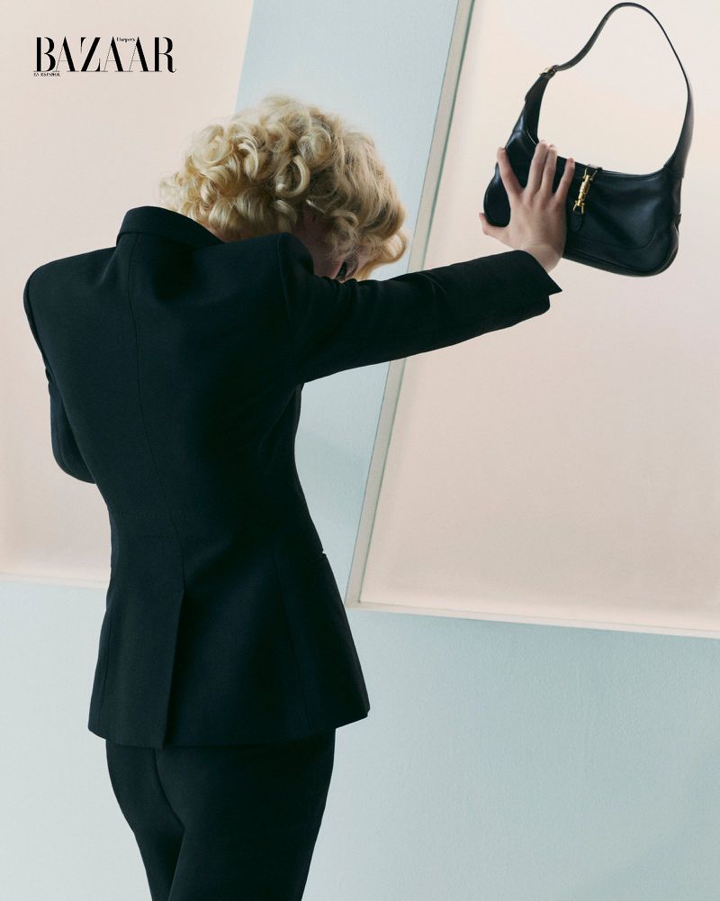 Fashion editorial shot of a person in a dark suit holding a black leather handbag in front of a light pastel background with graphic elements.