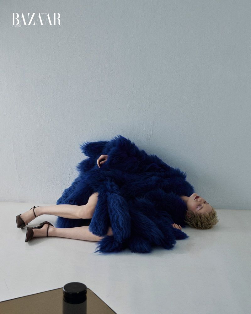 Fashion editorial shot of a blonde model reclining in a large blue furry coat, with dark brown heeled shoes, on a plain background.