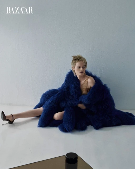 Fashion editorial shot of a woman in a blue faux fur coat and heels, posing against a light blue wall with the Bazaar logo visible.
