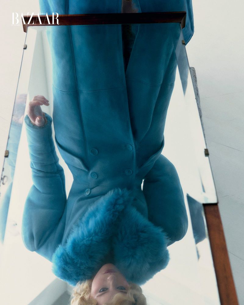 Fashion editorial shot from a low angle featuring a blonde model in a blue coat with fur trim reflecting in a mirror against a white background.