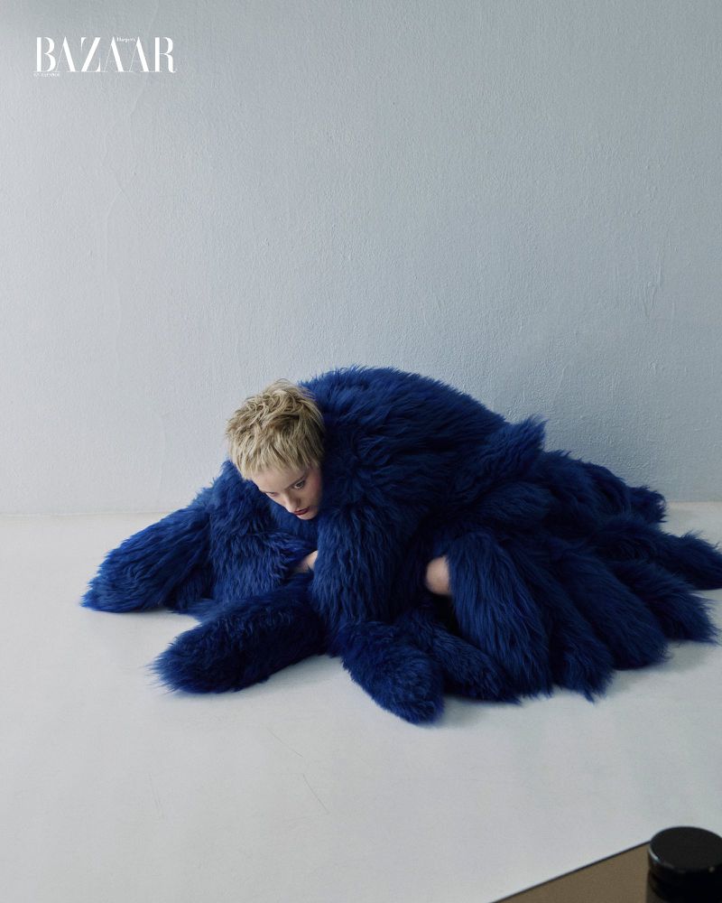 A fashion shot features a fair-skinned woman with short blonde hair partially submerged in a textured royal blue fur coat against a light blue background.
