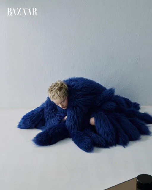 A fashion shot features a fair-skinned woman with short blonde hair partially submerged in a textured royal blue fur coat against a light blue background.