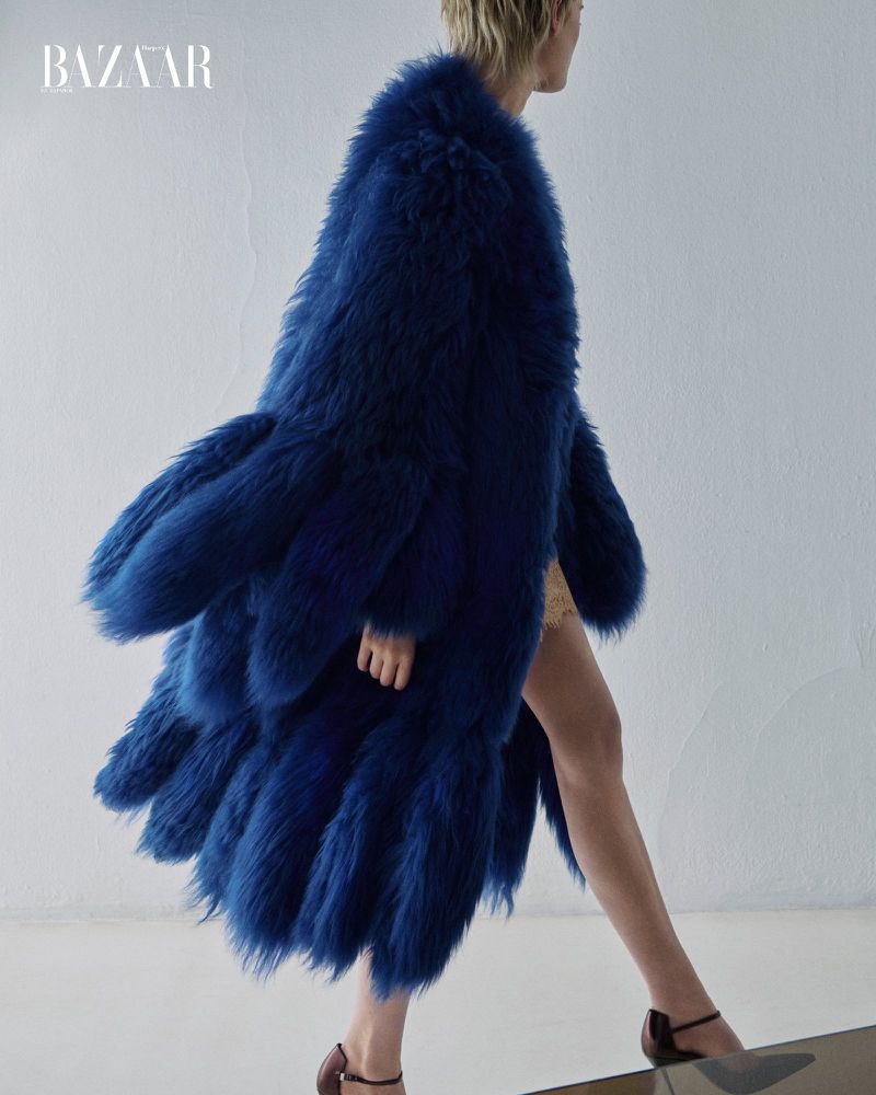 Model wearing a voluminous blue faux fur coat walking against a plain white studio wall for an editorial shoot.