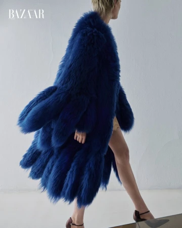 Model wearing a voluminous blue faux fur coat walking against a plain white studio wall for an editorial shoot.