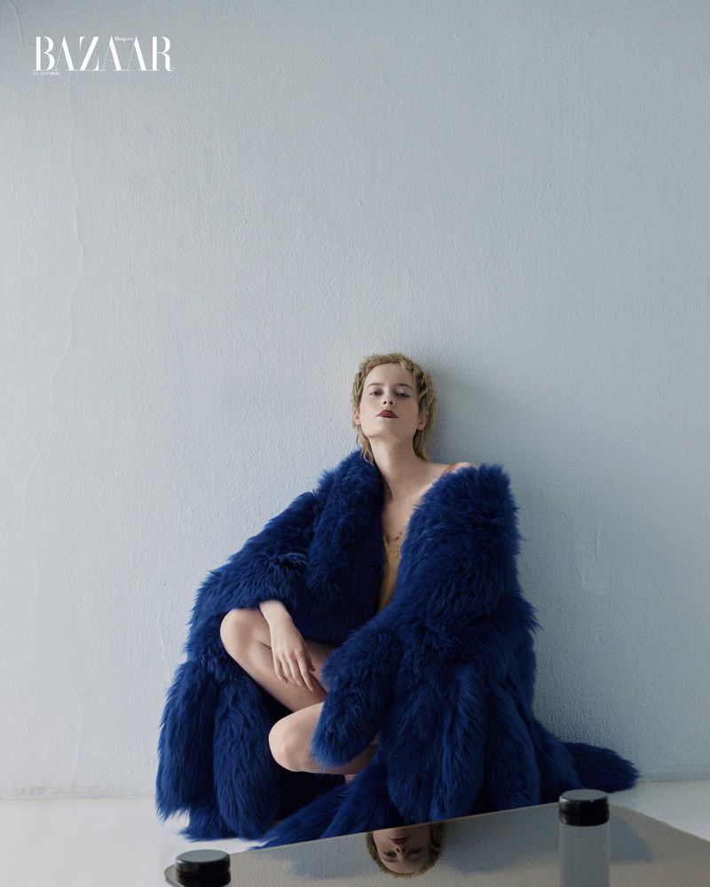 Fashion editorial shot of an adult woman sitting in a blue feather coat against a light grey wall with the Bazaar logo.