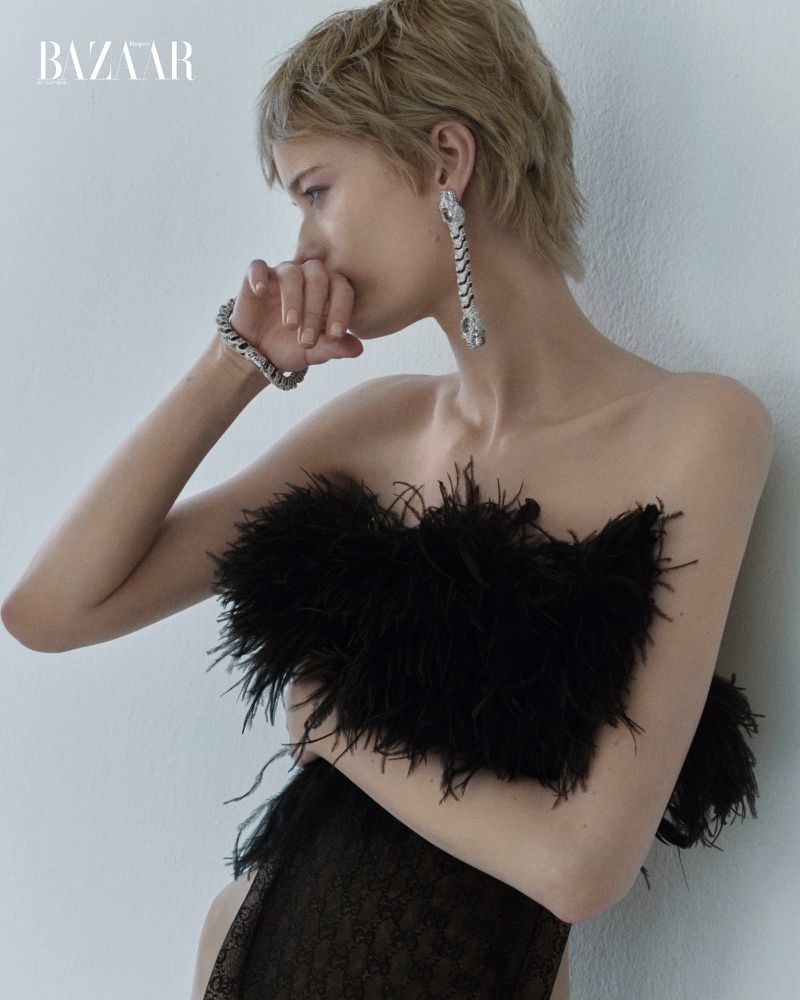 Portrait of a blonde woman in a feathered top and jewelry, gazing to the side in a studio setting. Bazaar logo visible in the upper left.