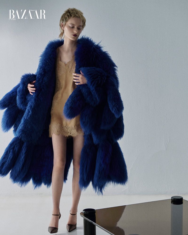Fashion editorial shot of a female model in a blue fur coat and beige dress, posing in a studio with dark heels and a glossy table.
