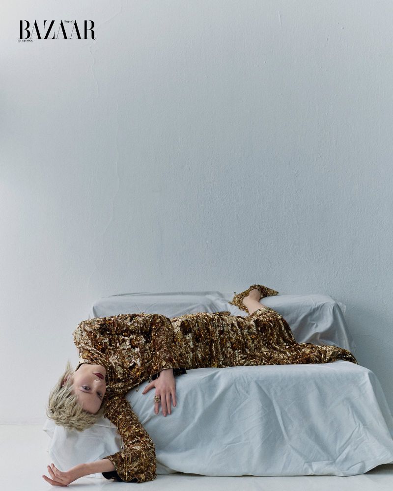 An adult female fashion model in an elegant dress lies on a sheet-covered sofa in a photo studio against a white wall with BAZAAR logo.