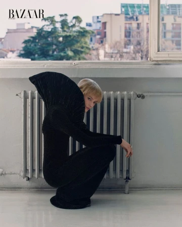 A blonde woman in a black dress poses inside next to a radiator and a window with city views for Bazaar magazine fashion editorial.