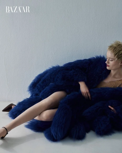 Fashion shot of a woman in a blue fur coat reclining on a white floor, legs crossed, dark heels, fair skin, and Bazaar logo visible.