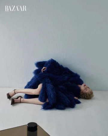 Model lying on the floor wearing a voluminous blue faux fur coat with high heels against a pale wall for Harper's Bazaar Mexico