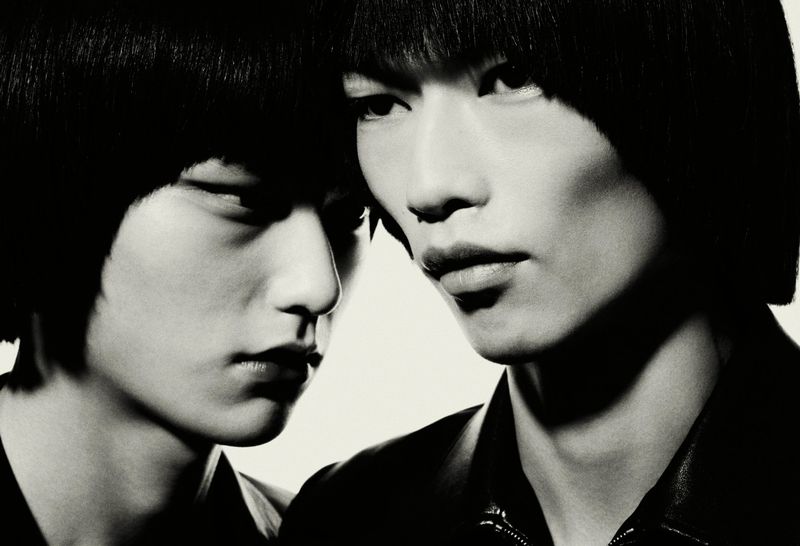 Striking black and white portrait of two Asian adult males with matching bowl haircuts, wearing dark jackets, against a white background.