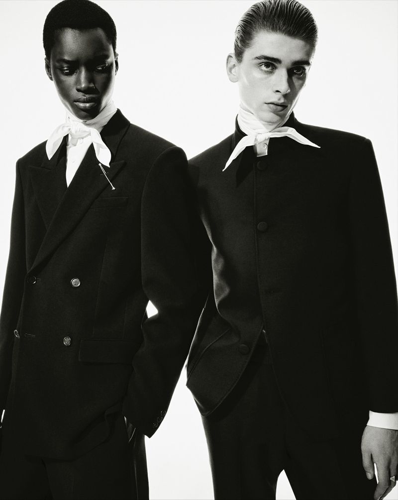 Black and white portrait of two stylish adults in black suits and white ascots, posing in a studio setting with a bright background.