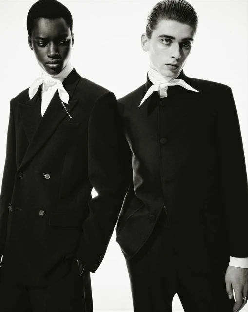 Black and white portrait of two stylish adults in black suits and white ascots, posing in a studio setting with a bright background.