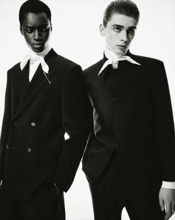 Black and white portrait of two stylish adults in black suits and white ascots, posing in a studio setting with a bright background.