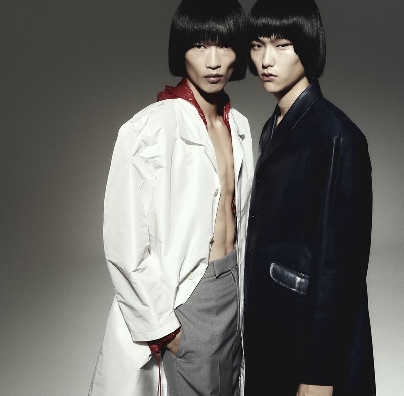 Two Asian men in contemporary fashion, one in a white trench coat and grey pants, the other in a black leather coat against a grey background.