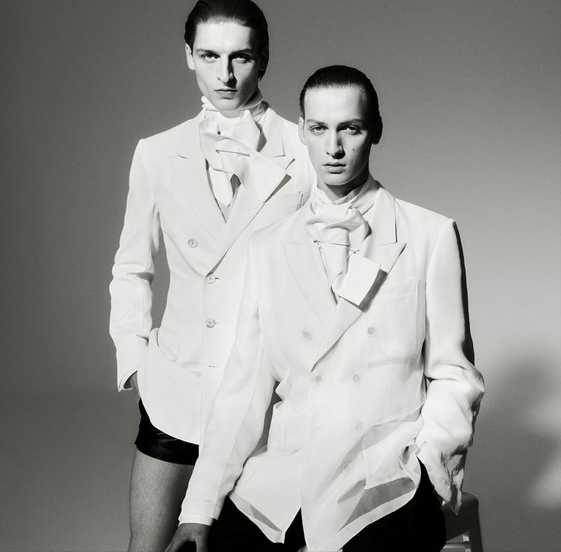 Black and white studio shot of two adult men in white double-breasted blazers, neck scarves, and dark shorts, posing indoors against gray backdrop.