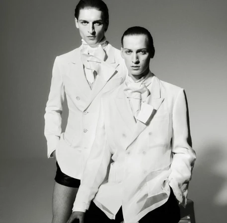 Black and white studio shot of two adult men in white double-breasted blazers, neck scarves, and dark shorts, posing indoors against gray backdrop.