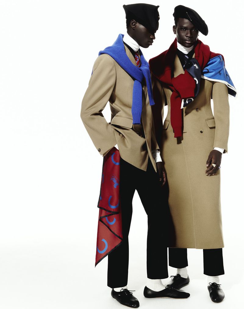 Two dark-skinned men model beige overcoats and black berets in a studio, accessorized with colorful scarves, set against a plain white backdrop.
